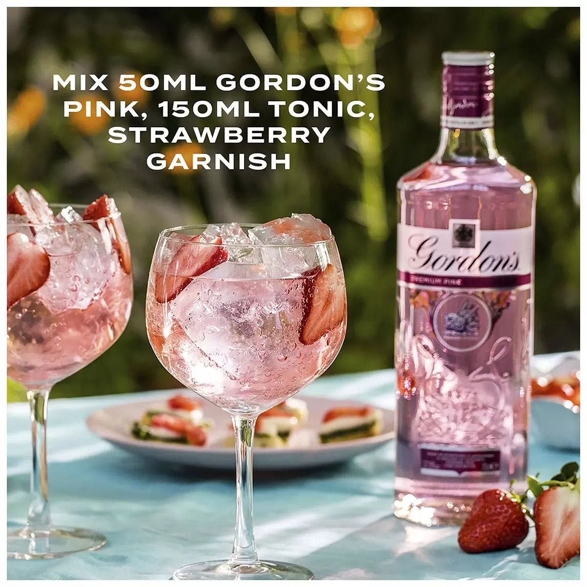 Gordon's Pink gin bottle with two glasses of pink cocktail and strawberries on a table outdoors.