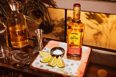 Bottle of Jose Cuervo on a tray with limes and salt, against a reflective gold surface.