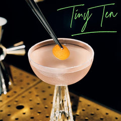 Cocktail in a pink glass with a lemon twist on a black background, featuring 'Tiny Tea' text.