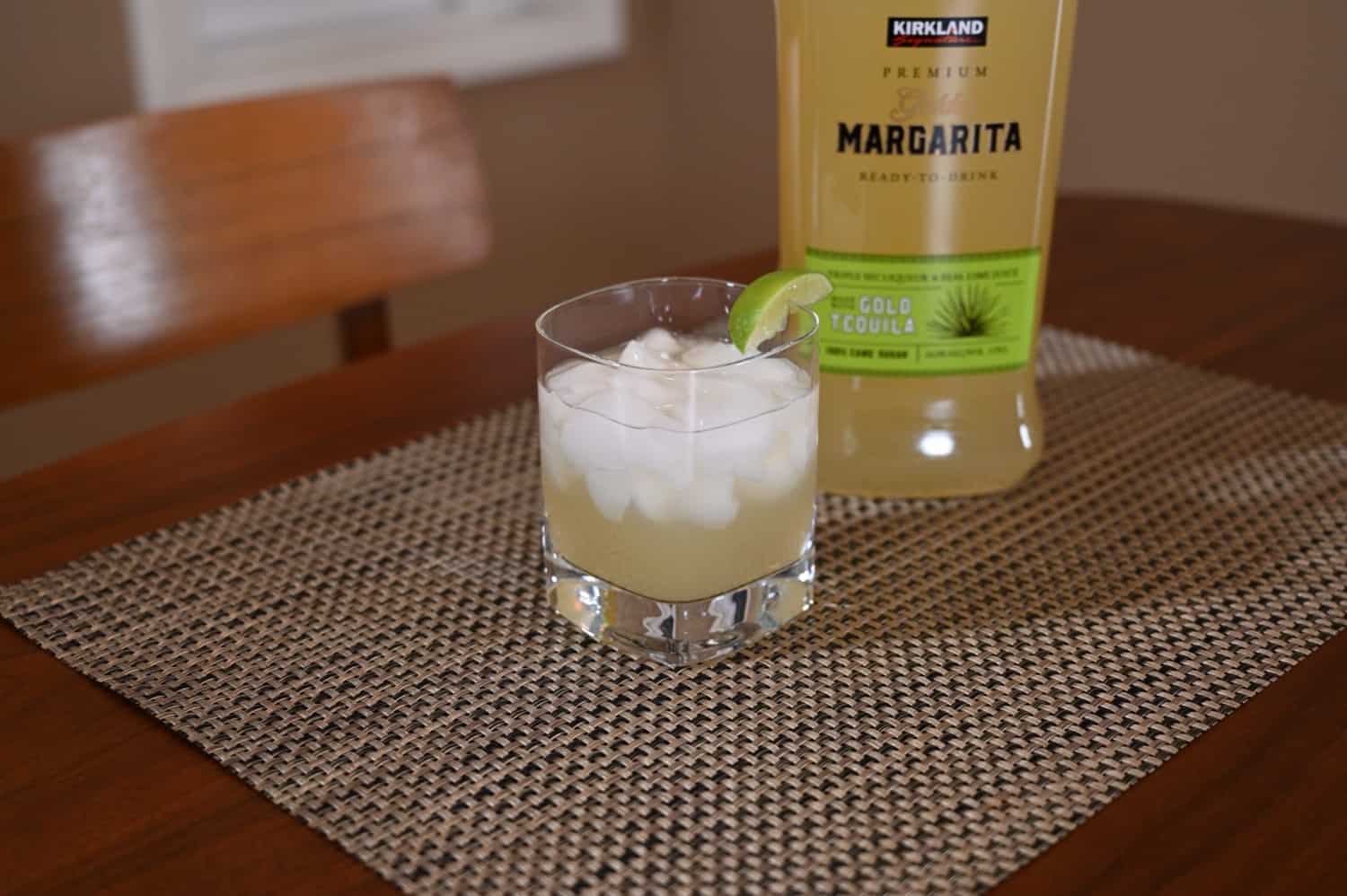 Glass of margarita with lime wedge next to a bottle of Kirkland margarita on a textured surface.