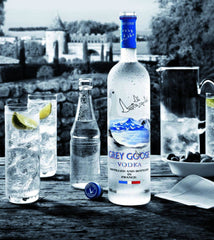 Bottle of Grey Goose vodka on a table with glasses and a pitcher in an outdoor setting.