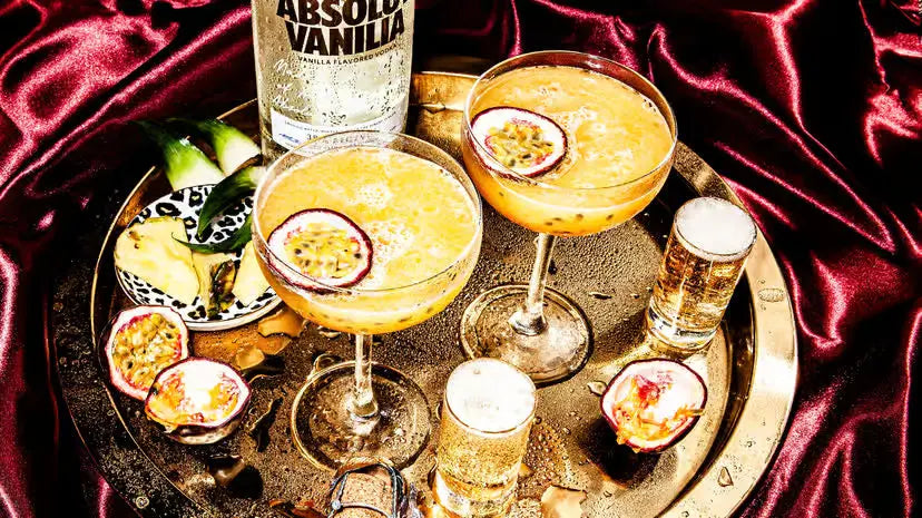 Two cocktails with passion fruit garnishes on a gold tray with an Absolut Vanilla bottle, surrounded by red fabric.
