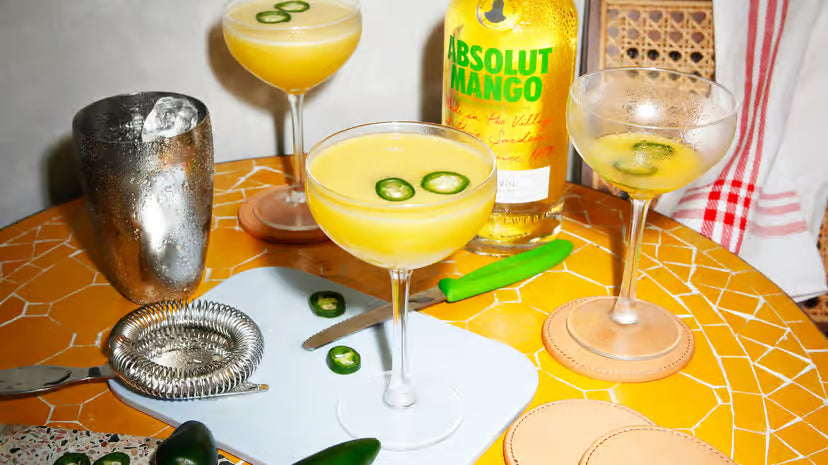 Cocktails with jalapeño slices, a bottle of Absolut Mango, and bar tools on a yellow patterned table.