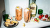Two cocktails with ice and garnishes, a bottle of Absolut Vanilla vodka, and snacks on a white surface.