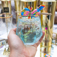 Hand holding a clear glass with blue liquid, gold rim, and colorful drink stirrer against a blurred background.