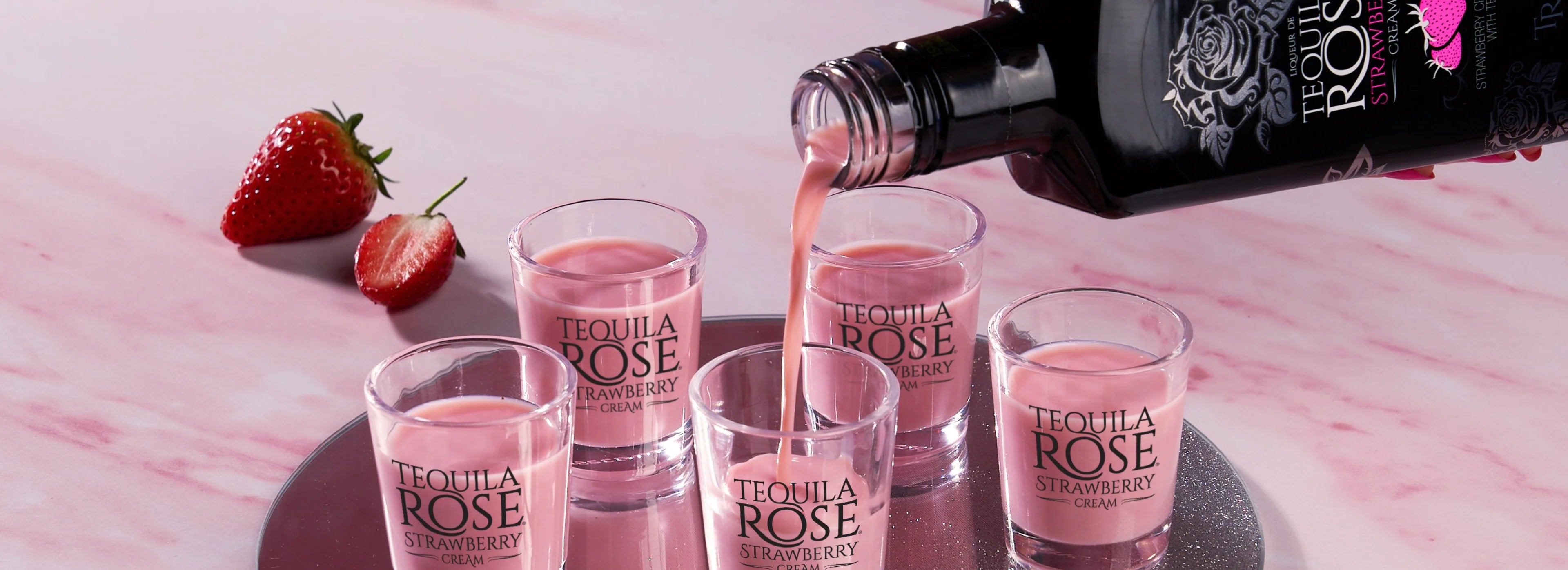 Tequila Rose Strawberry being poured into glasses with a strawberry on a pink marble surface