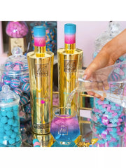 Gold bottles with colorful caps being poured into a glass filled with blue and pink liquid, surrounded by candy jars.