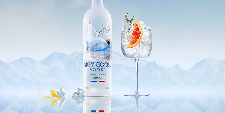 Grey Goose vodka bottle with a glass of vodka and orange slice against a mountainous background