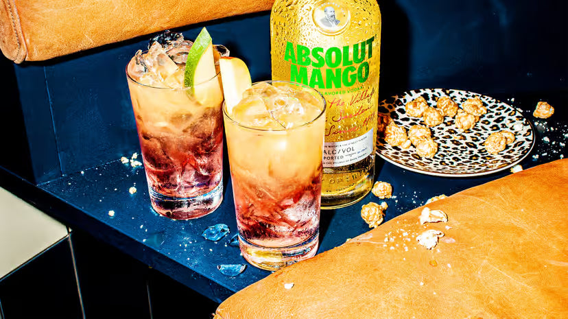 Two cocktails with ice and a lime garnish next to an Absolut Mango bottle on a bar counter.